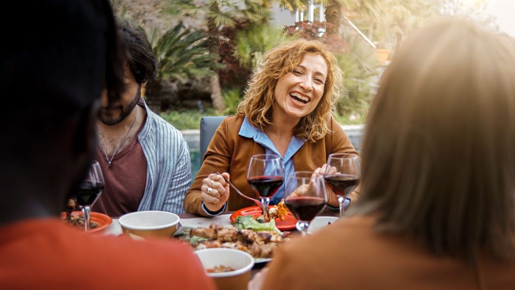 Positive forty years old woman smiles, eats, drinks and talks with friends eating at the bbq table in the farmhouse courtyard sitting at the brunch table - food and drink people life style concept